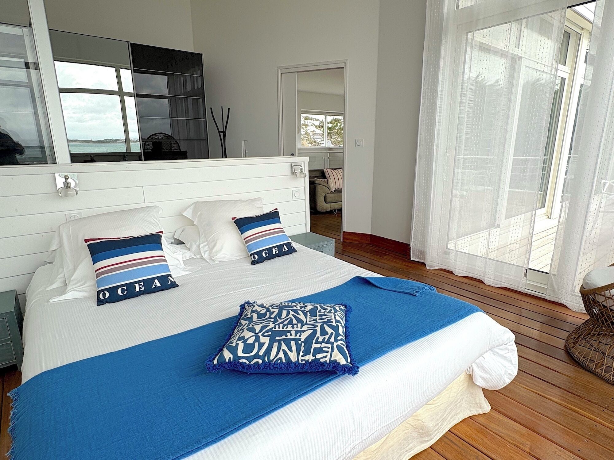 A bedroom with a large bed, white headboard, and a view of the outdoors through a window with sheer curtains.