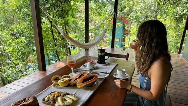 Une femme est assise à une table en bois, devant un paysage verdoyant, avec un hamac à proximité et un repas composé de pain, de fruits et d’une boisson.