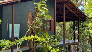 Une cabane rustique en bois, avec un toit de chaume, entourée d’une végétation luxuriante et d’un grand tronc d’arbre.