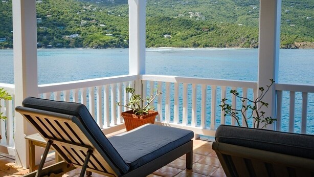 A covered balcony with two lounge chairs and a potted plant, overlooking a body of water and green hills. - 3 Mins to Hull Bay | Dining | Quiet Retreat (St. Thomas)