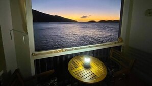 A room with a large window overlooking a body of water at sunset, a small round table with a light, and a chair. - Serene Escape + 3 Mins to Beach + Dining (St. Thomas)