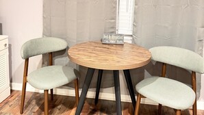 Una stanza con un tavolo rotondo in legno e quattro sedie rivestite di verde chiaro. Un cartello sul tavolo recita “Bed & Breakfast You Make Both”. La stanza ha un soffitto in legno e una finestra con persiane.