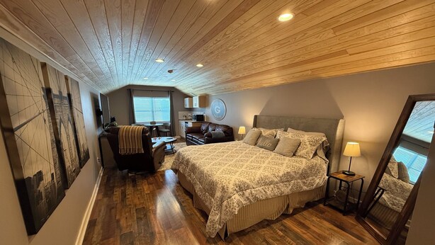 Una camera da letto con soffitto in legno, un letto con lenzuola fantasia, una scrivania, una sedia, un divano, una TV e una finestra con persiane.