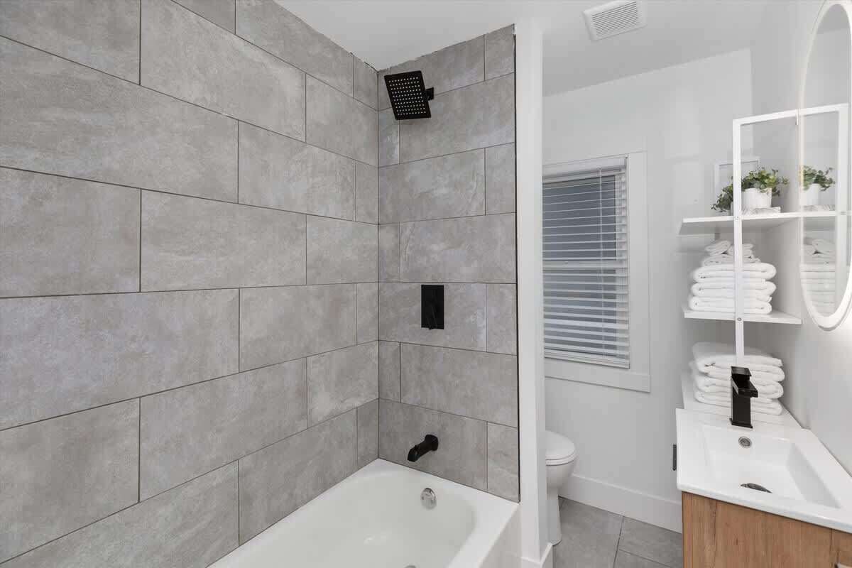 A modern bathroom with grey tiles, a white bathtub, a toilet, and a white sink with a mirror.