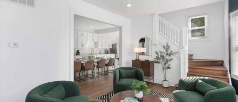 A modern living room with a round coffee table, two green armchairs, a black and white rug, and a staircase leading to an upper level.