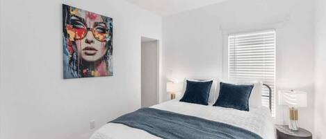A bedroom with a bed, a painting on the wall, a window with blinds, and bedside lamps.