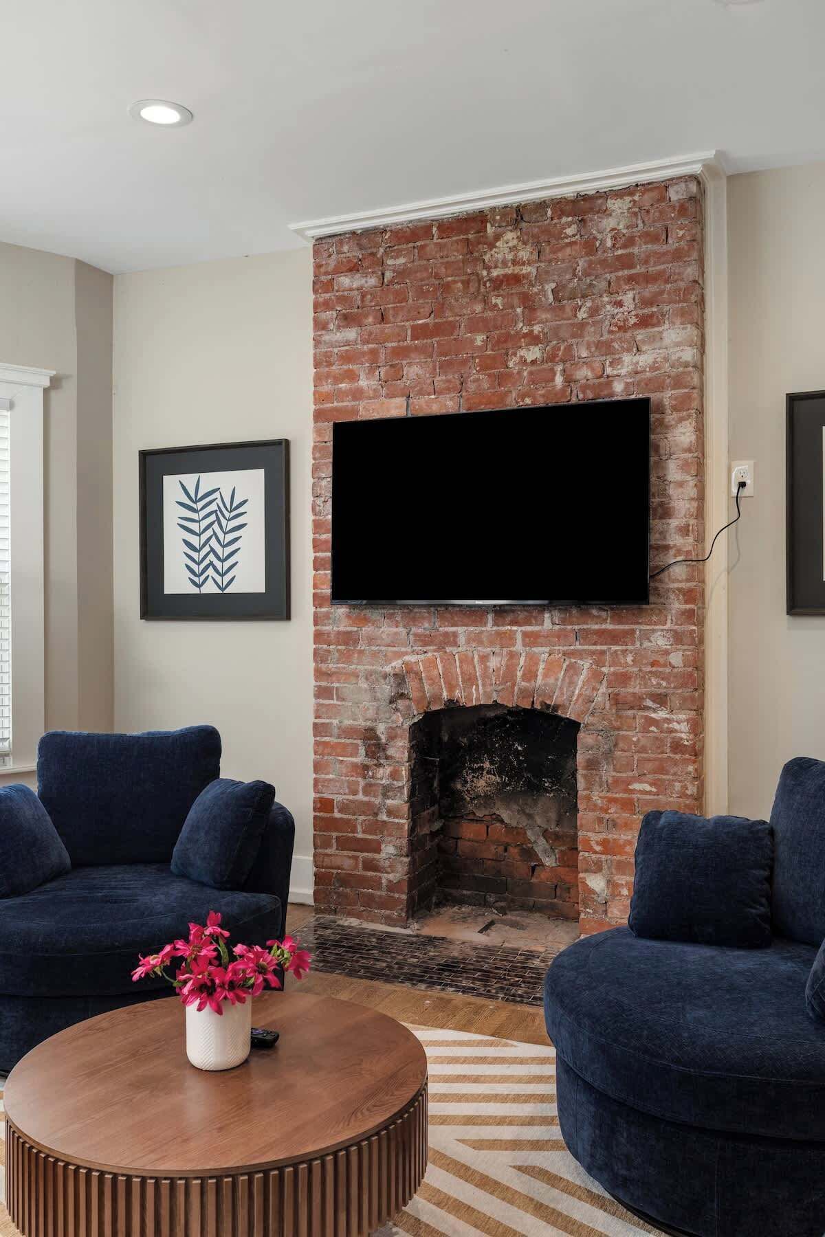 A living room with a brick fireplace, two dark sofas, a round coffee table with a vase of flowers, and a flat-screen TV mounted above the fireplace.