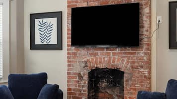 A living room with a brick fireplace, two dark sofas, a round coffee table with a vase of flowers, and a flat-screen TV mounted above the fireplace.