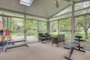 A gym with exercise equipment, wicker chairs, and large windows overlooking a garden.