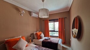 A hotel room with two beds, a large window with orange curtains, a hanging light fixture, and a wall-mounted mirror. - Cosy apartment in the heart of the medina (Marrakech)