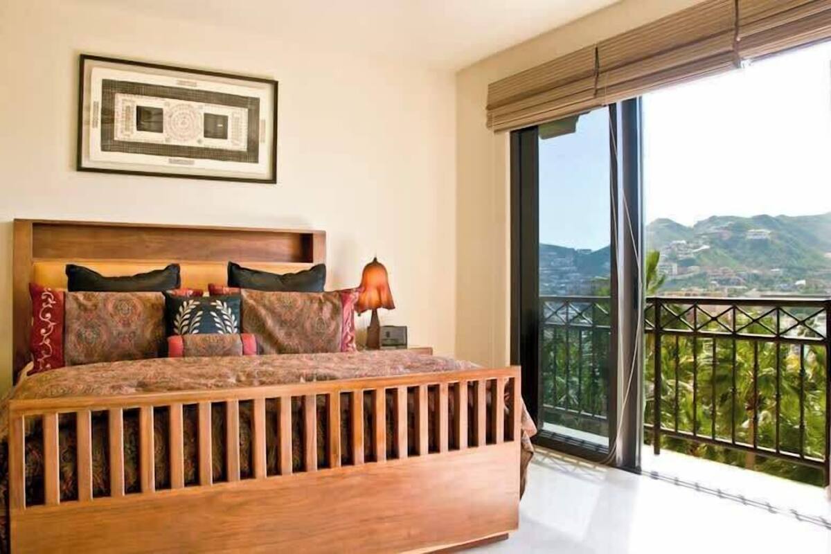 A bedroom with a large bed, a nightstand, a framed picture above the bed, and a balcony with a view of the hills.