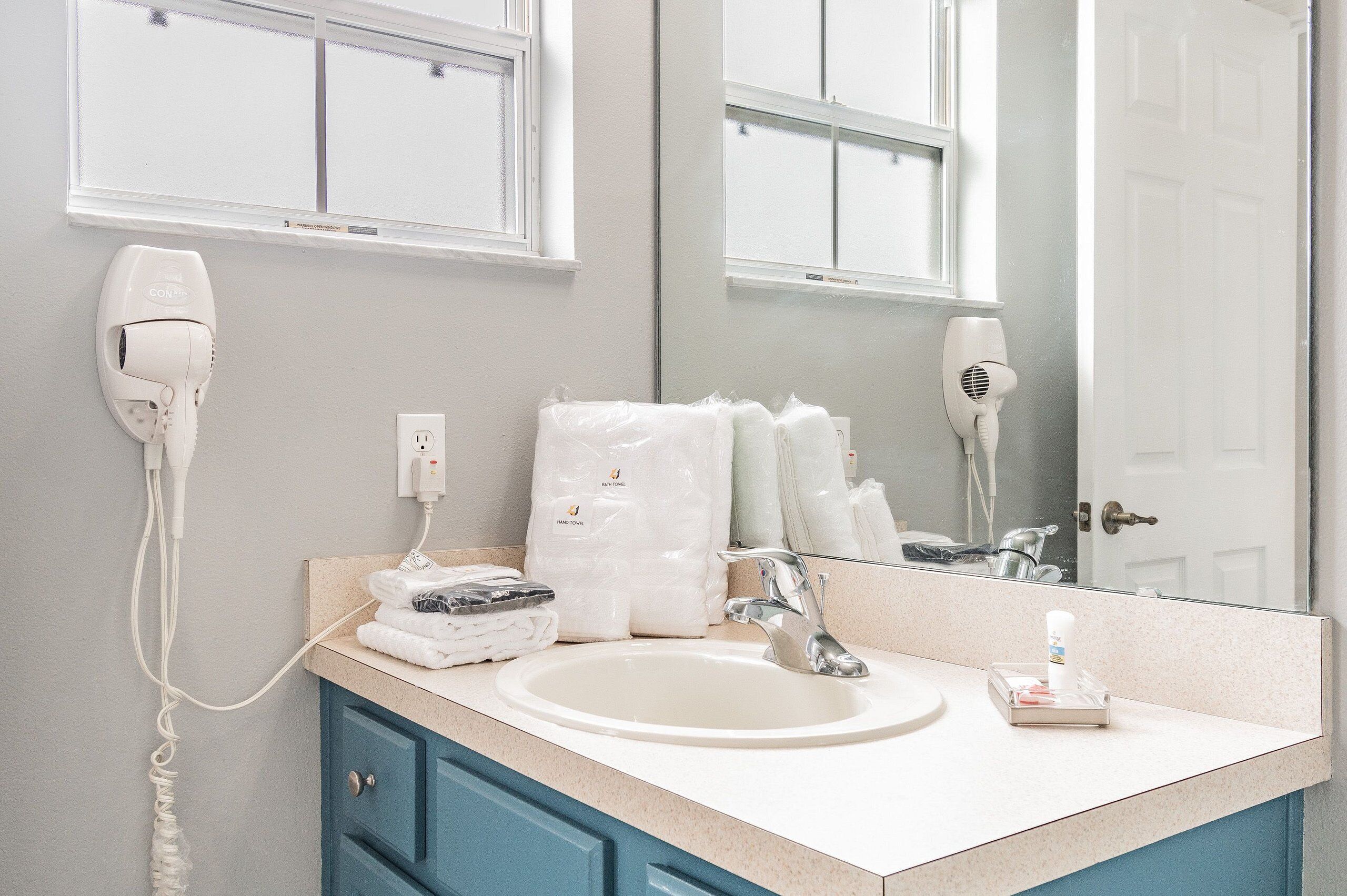 A bathroom with a sink, two hair dryers, a mirror, and a towel dispenser.