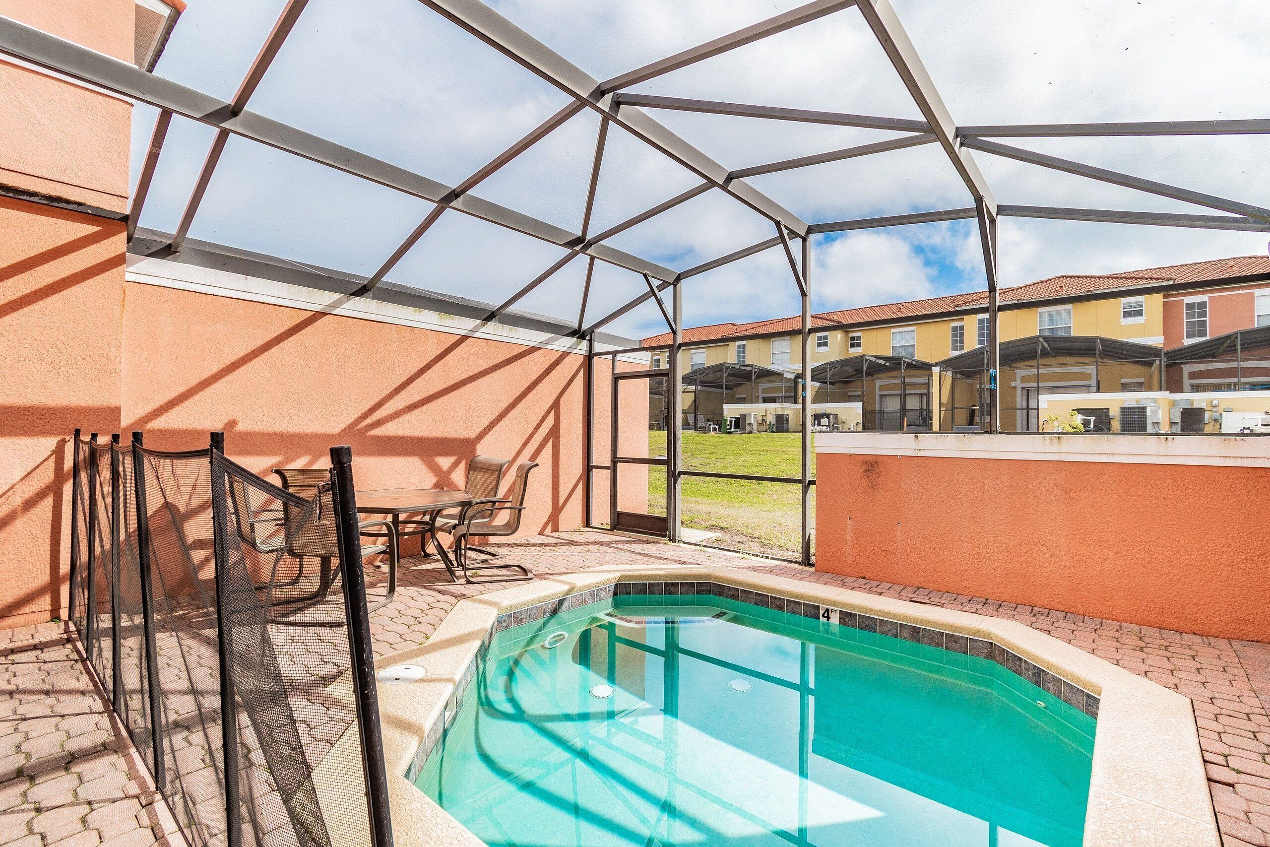 A covered outdoor swimming pool with a patio area, metal fencing, and a view of neighboring buildings.