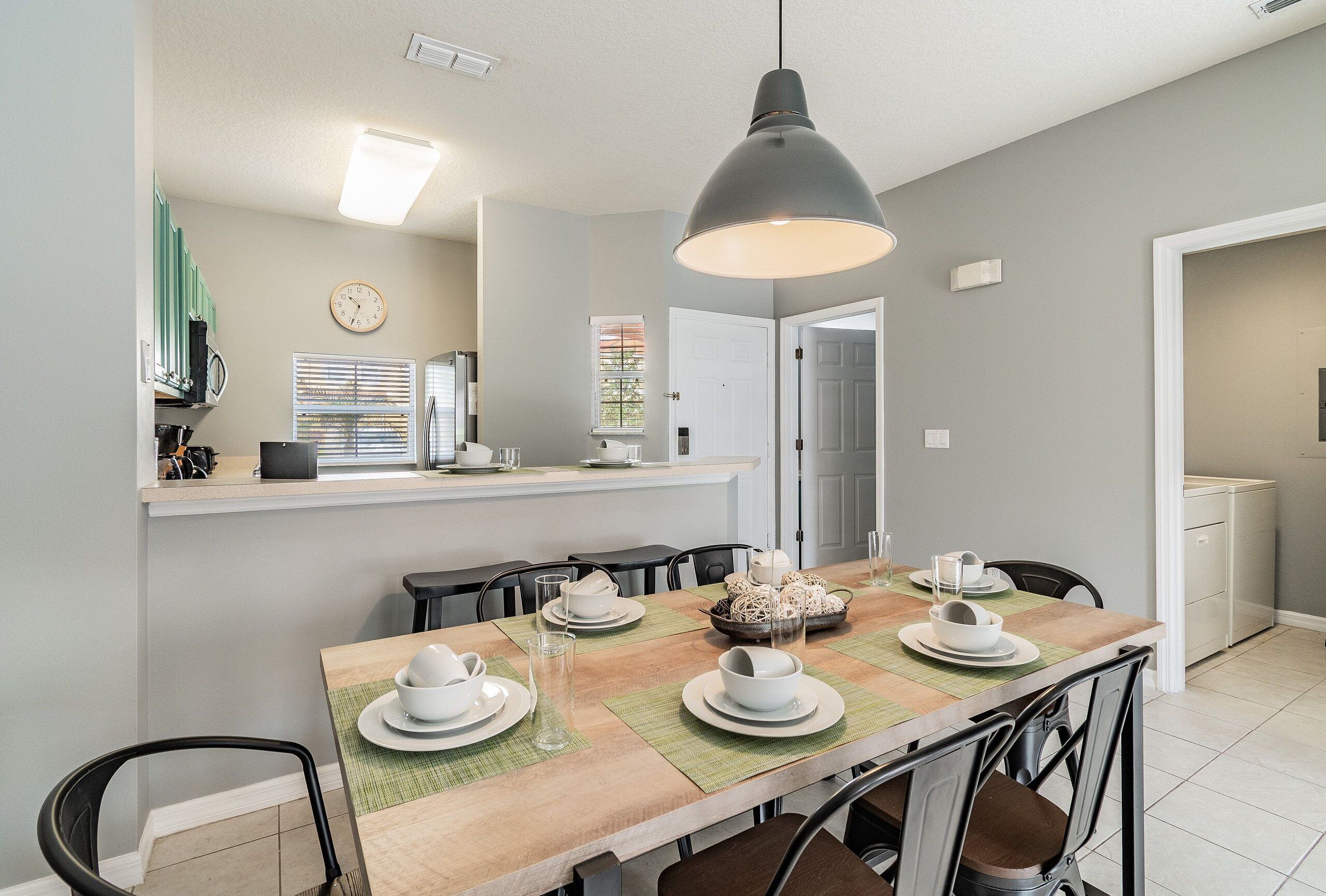 A dining area with a wooden table set for four, surrounded by chairs. A large pendant light hangs above the table.