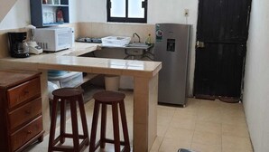 A compact kitchen with a refrigerator, microwave, and sink. There are two stools and a wooden table. A black door and a window are visible. - Bungalow Turrent, 5 minutes from Playa Mocambo, Boca del Rio Veracruz (Veracruz)