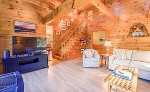 A log cabin interior with a TV, sofa, armchair, wooden staircase, and a bookshelf.