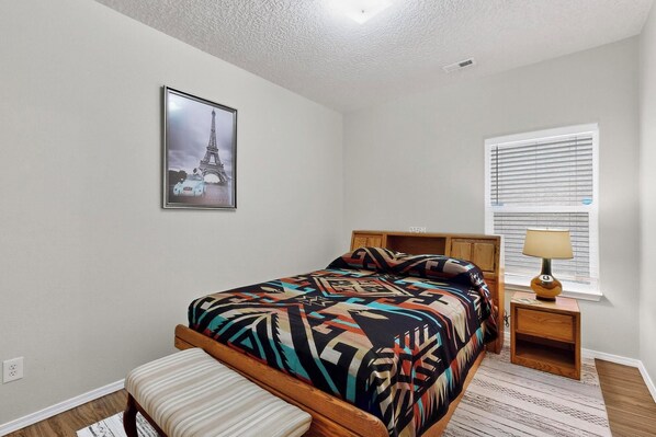 A bedroom with a bed, a nightstand, a lamp, a window with blinds, and a picture of the Eiffel Tower on the wall.