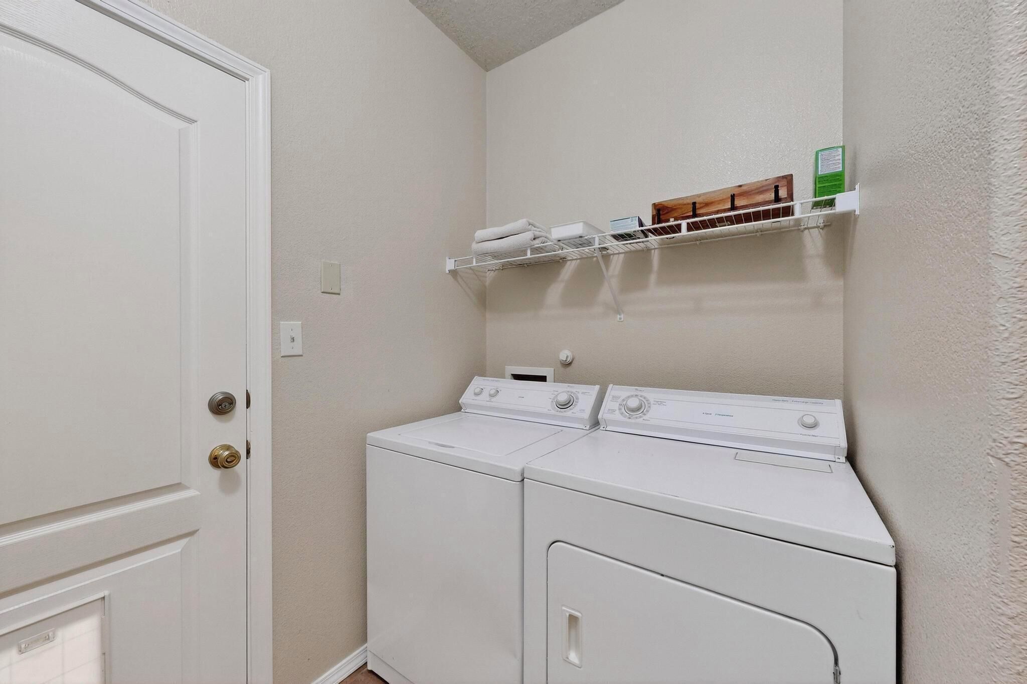 A laundry room with two white front-loading washers and dryers, a white door, and a shelf above the dryers holding folded towels and a green box.