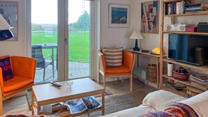 Ein Wohnzimmer mit einer Couch, einem Sessel und einem Tisch mit Fernbedienung. Ein Bücherregal mit Büchern und ein Fernseher sind ebenfalls vorhanden.