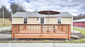 A single-story building with a wooden deck, a covered patio area, and a red barn in the background. - NYC Escape | Pocono Cabin & Suite (Pleasant Mount)