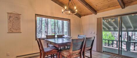 A dining area with a wooden table and chairs, large windows with a forest view, and a hanging light fixture.
