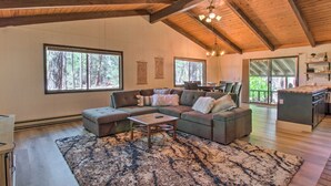 A spacious living area with a large sectional sofa, a coffee table, and a rug. There are large windows offering views of the forest.