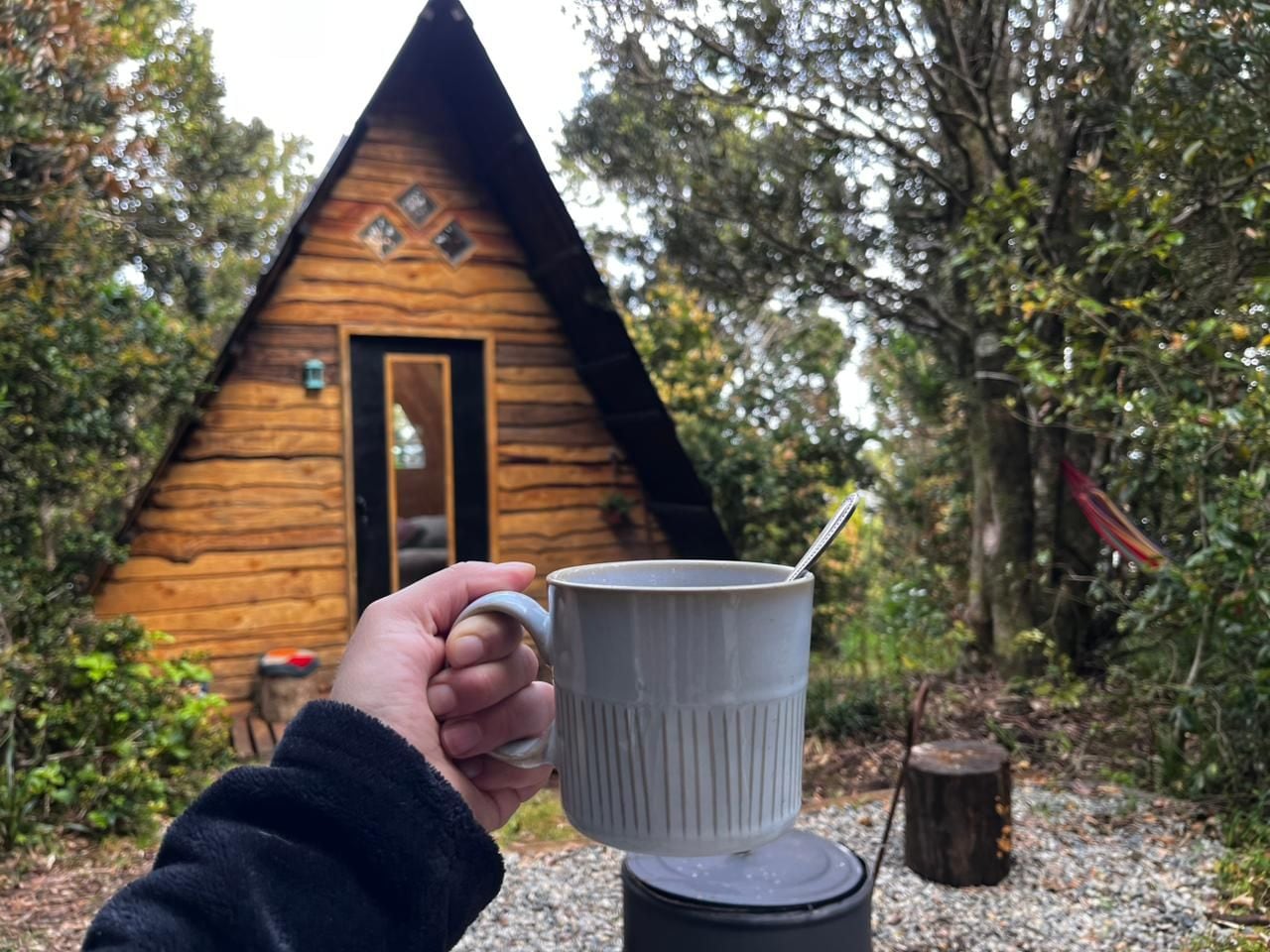 A hand holding a white mug with a spoon, in front of a wooden cabin with a door and windows.