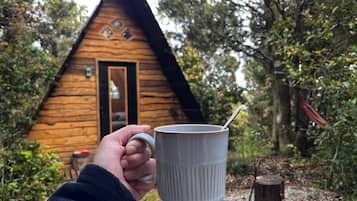 A hand holding a white mug with a spoon, in front of a wooden cabin with a door and windows.