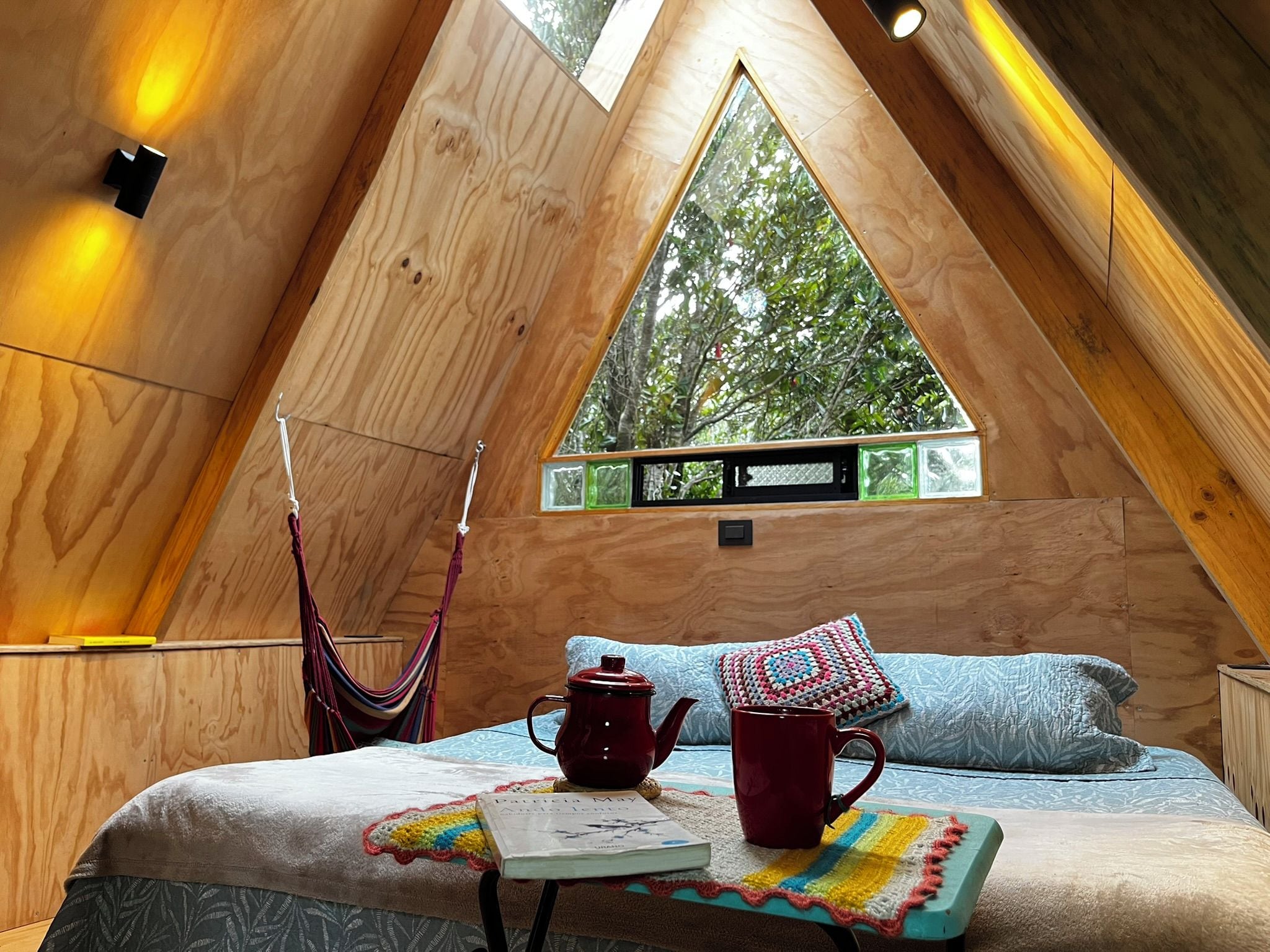 A cozy room with a sloped ceiling, a hammock, a bed with a colorful blanket, a red kettle, and a window with a view of trees.