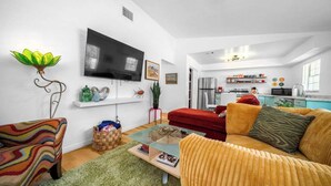 A modern living room with a flat-screen TV, colorful furniture, and a kitchen in the background.