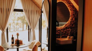 Um quarto com uma mesa redonda, cadeiras, uma janela com cortinas e um arco de pedra que dá para o banheiro.