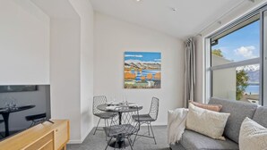 Een moderne woonkamer met een grijze bank, een houten salontafel en een eethoek met een ronde tafel en draadstoelen. Op de muur hangt een schilderij van een landschap, en er is een groot raam met uitzicht op een waterpartij.