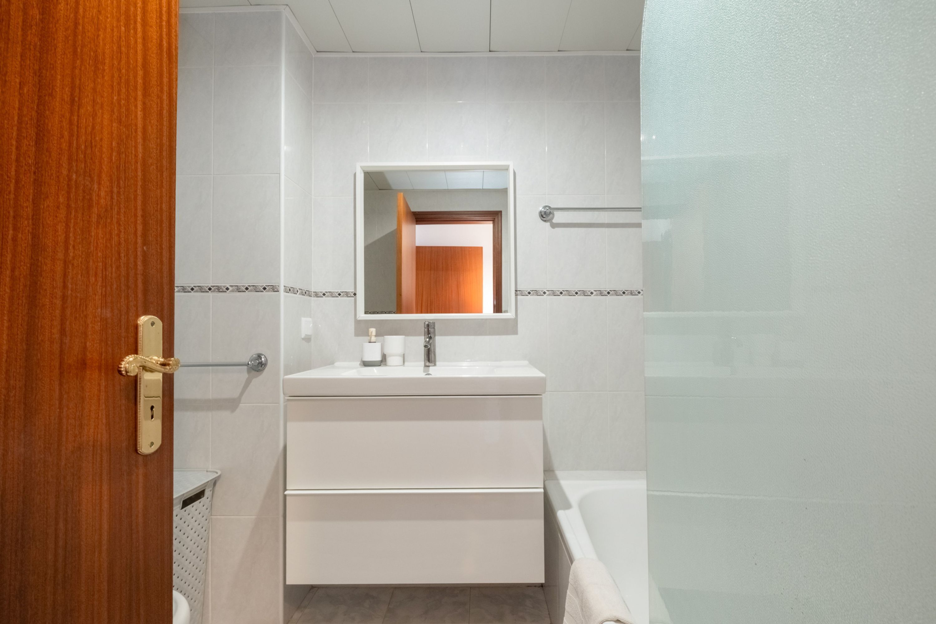 A bathroom with a white sink, mirror, and bathtub. There is a towel rack above the sink and a door with a golden handle.