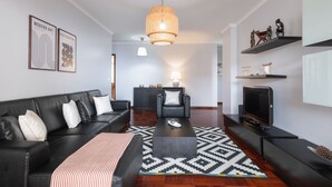 Una sala de estar moderna con un sofá de cuero negro, una mesa de centro y un televisor sobre un mueble bajo. Hay estantes montados en la pared con objetos decorativos y un colgante.