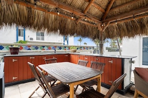 Un espacio al aire libre techado con mesa y sillas de madera, techo de paja y vista a un edificio con tejas de colores.