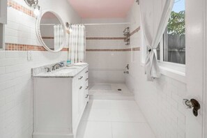 Un baño con paredes de azulejos blancos, un lavabo de mármol, una ducha y una ventana con cortinas.