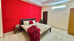 Een slaapkamer met een rode wand, een bed met donker hoofdeinde, een nachtkastje, een houten garderobe en een plafondventilator.
