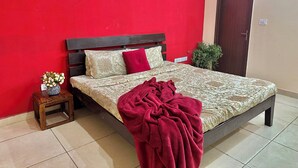 A bedroom with a red wall, a bed with a patterned bedspread, a wooden headboard, and a small bedside table with a vase of flowers.