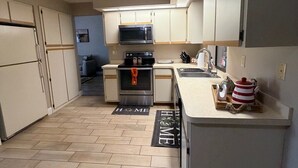 Een compacte keuken met een magnetron, oven en spoelbak. Er is een klein aanrecht met een waterkoker en een dienblad. De vloer heeft een 'Home' mat.