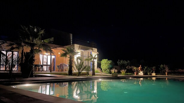A nighttime view of a hotel with a swimming pool, outdoor seating, and palm trees. - LaPerleD'argan (Essaouira)