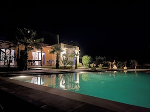 A nighttime view of a hotel with a swimming pool, outdoor seating, and palm trees.