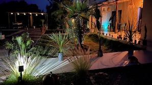 A well-lit outdoor area with a pathway, potted plants, and a building with a stone facade. - LaPerleD'argan (Essaouira)