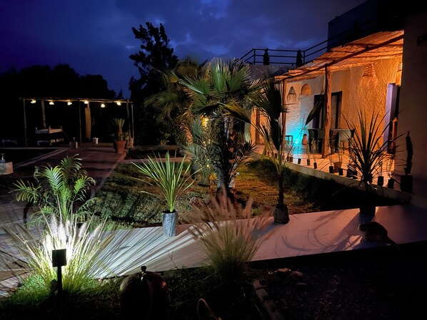 A well-lit outdoor area with a pathway, potted plants, and a building with a stone facade.
