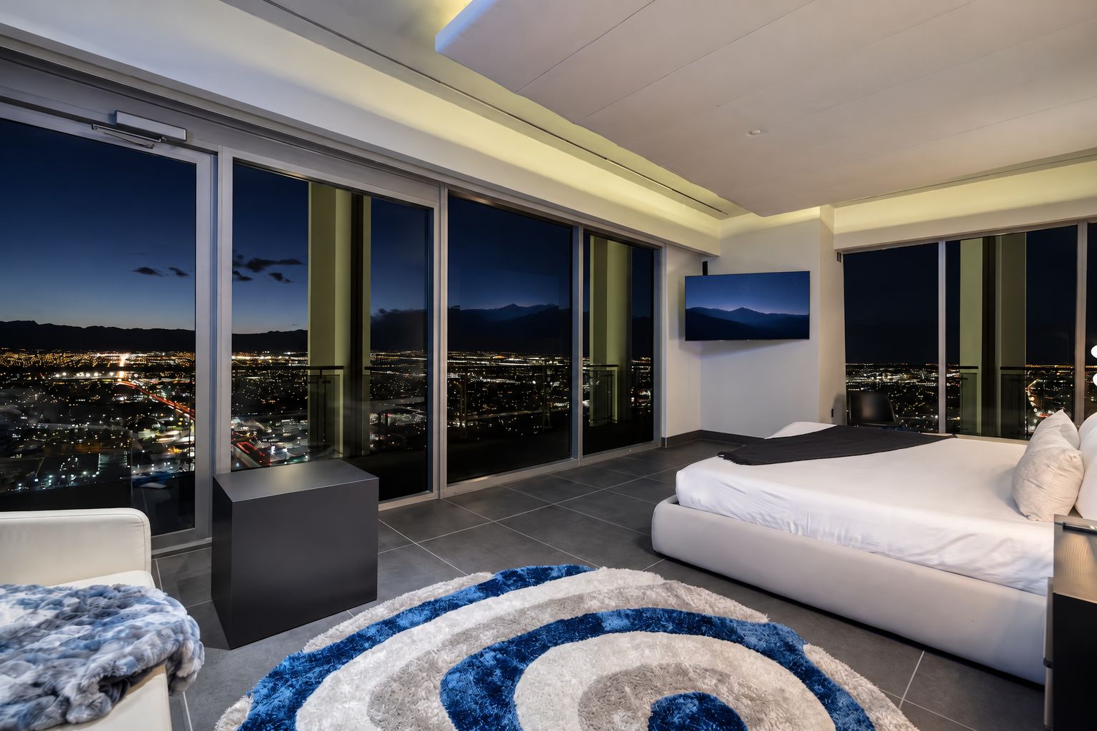 A modern hotel room with a large bed, a sofa, a TV, and a city view through floor-to-ceiling windows.