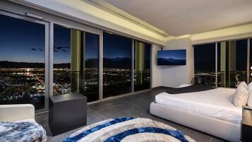 A modern hotel room with a large bed, a sofa, a TV, and a city view through floor-to-ceiling windows.