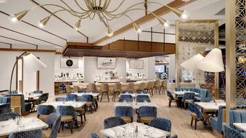 A restaurant with a modern interior, featuring blue upholstered chairs, white tables, and a bar area with a decorative wall.