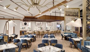 Un restaurant amb un interior modern, amb caires tapissades de blau, taules blanques i una zona de bar amb una paret decorativa.