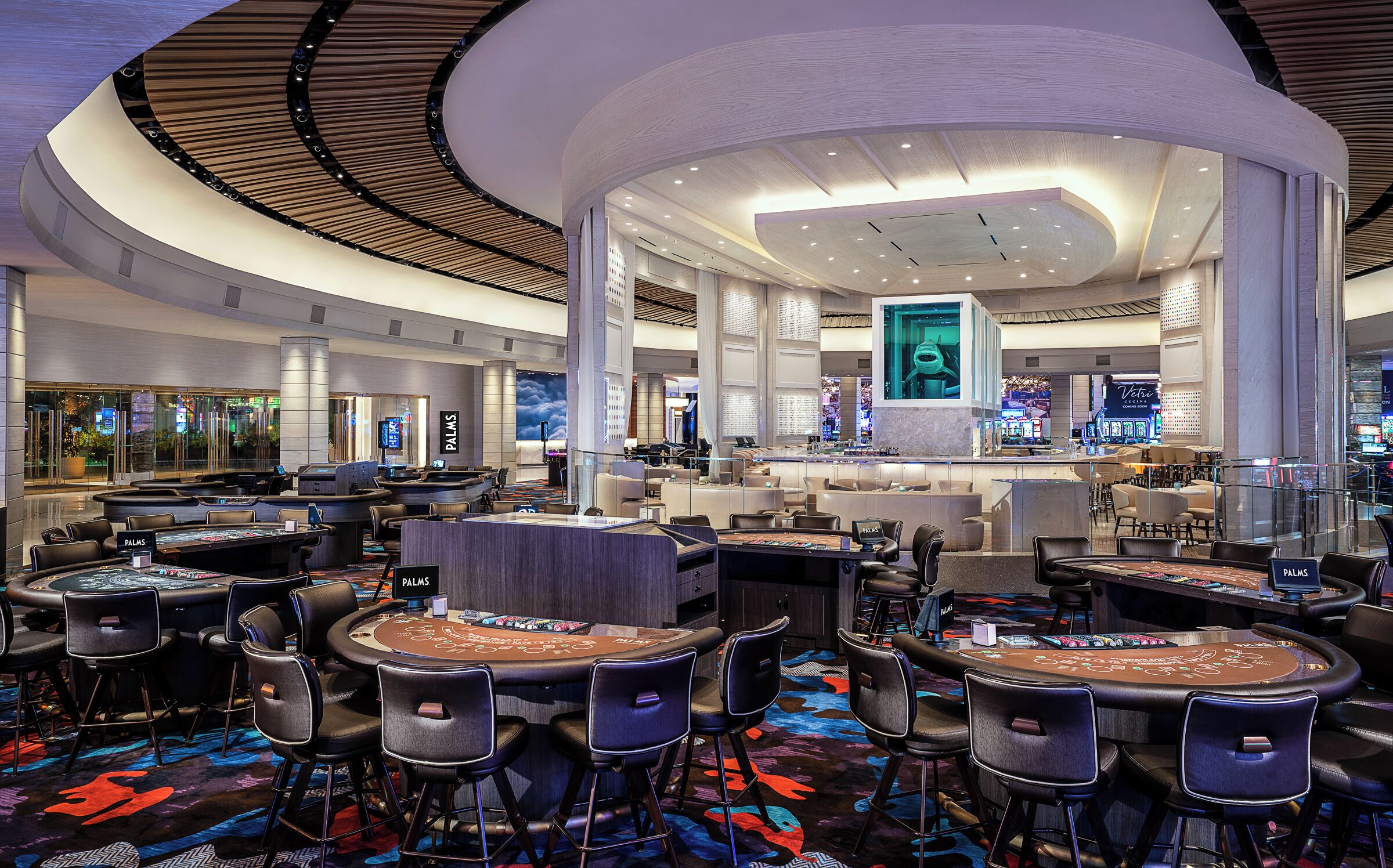 A modern casino interior with multiple blackjack tables, comfortable seating, and a large digital screen displaying a person.