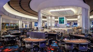 A modern casino interior with multiple blackjack tables, comfortable seating, and a large digital screen displaying a person.