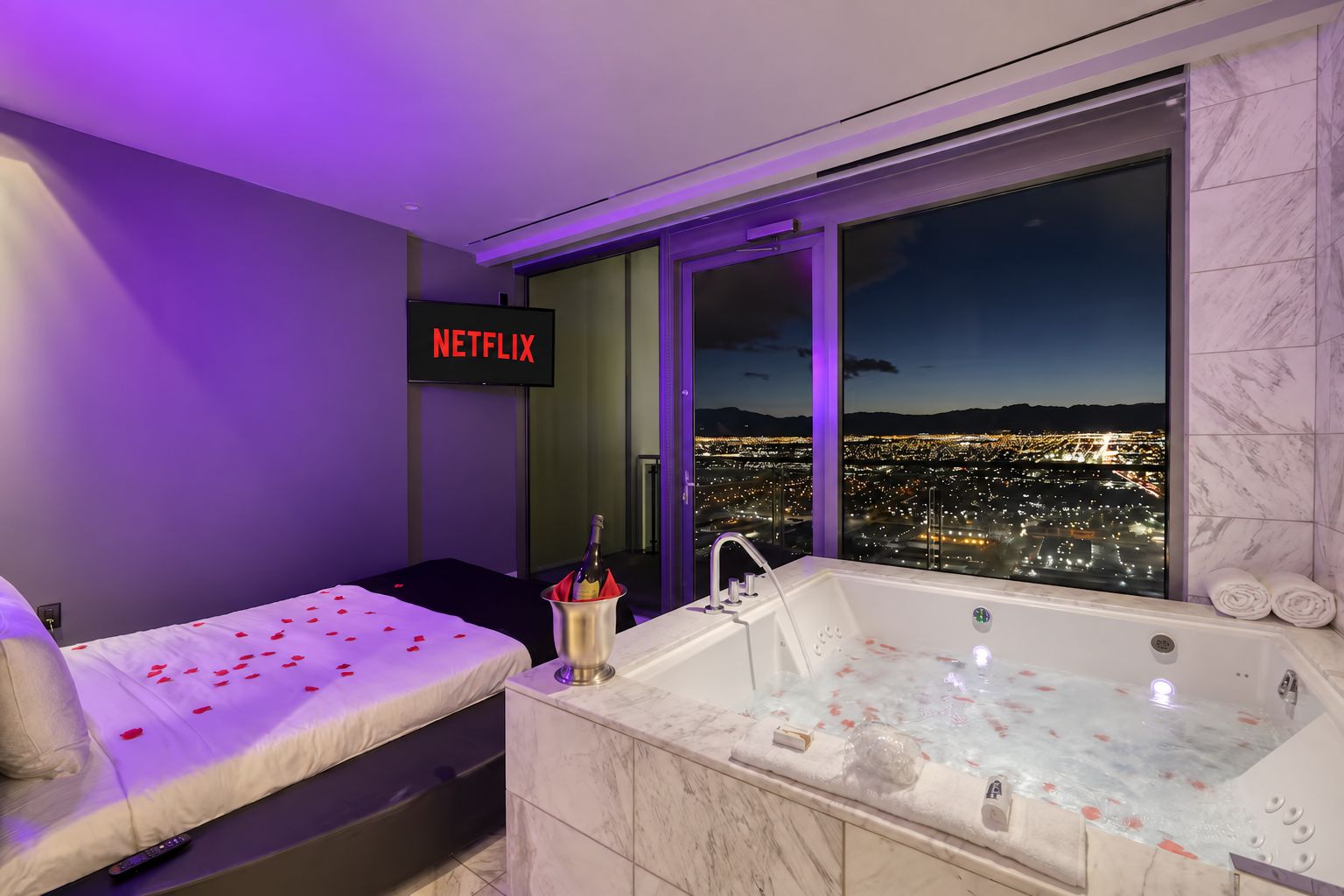 A hotel room with a bed, a large window with a city view, a bathtub, and a sign that says 'NETFLIX'.